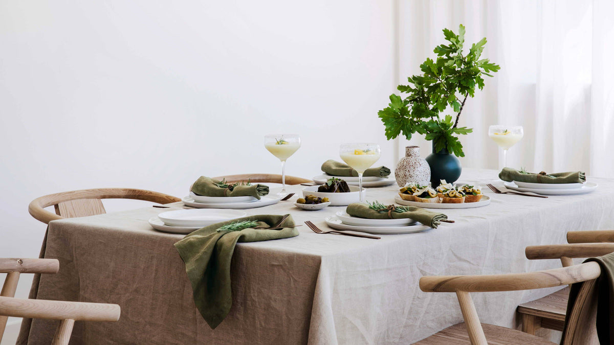 Dining table set with plates, napkins, and a vase with greenery in a bright room.