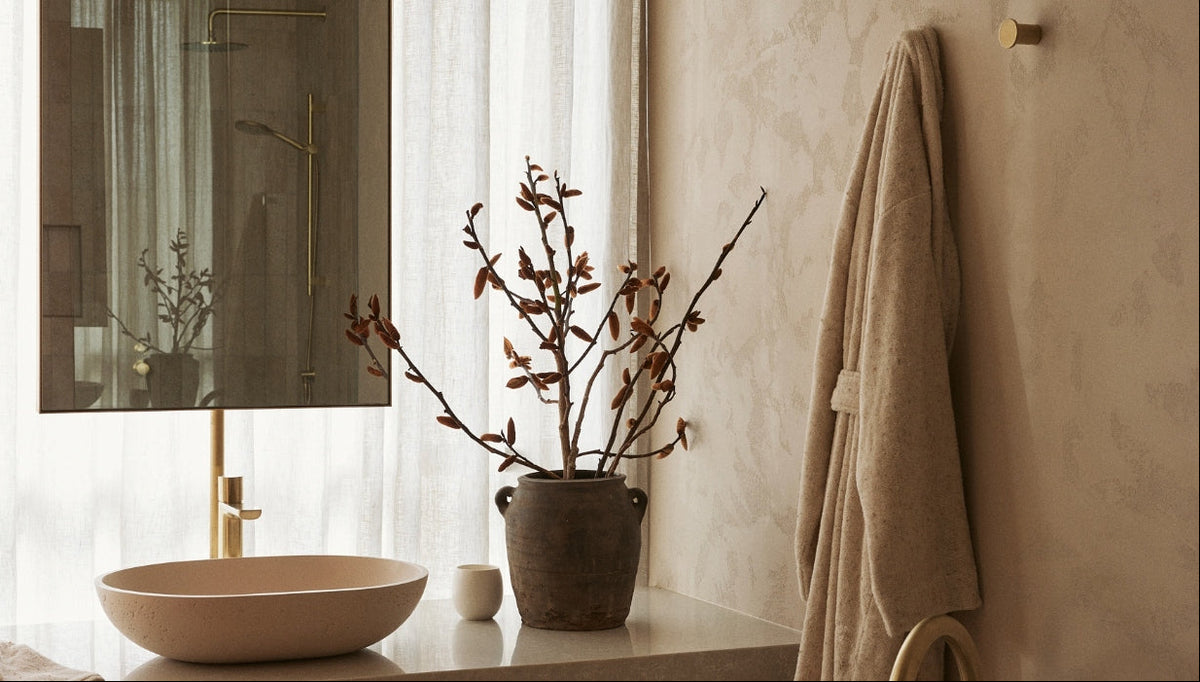 A beige bathroom with a round sink, a potted plant with brown leaves, a cup, a wall mirror, and a beige robe hanging on the wall. Light streams through sheer curtains behind the decor.