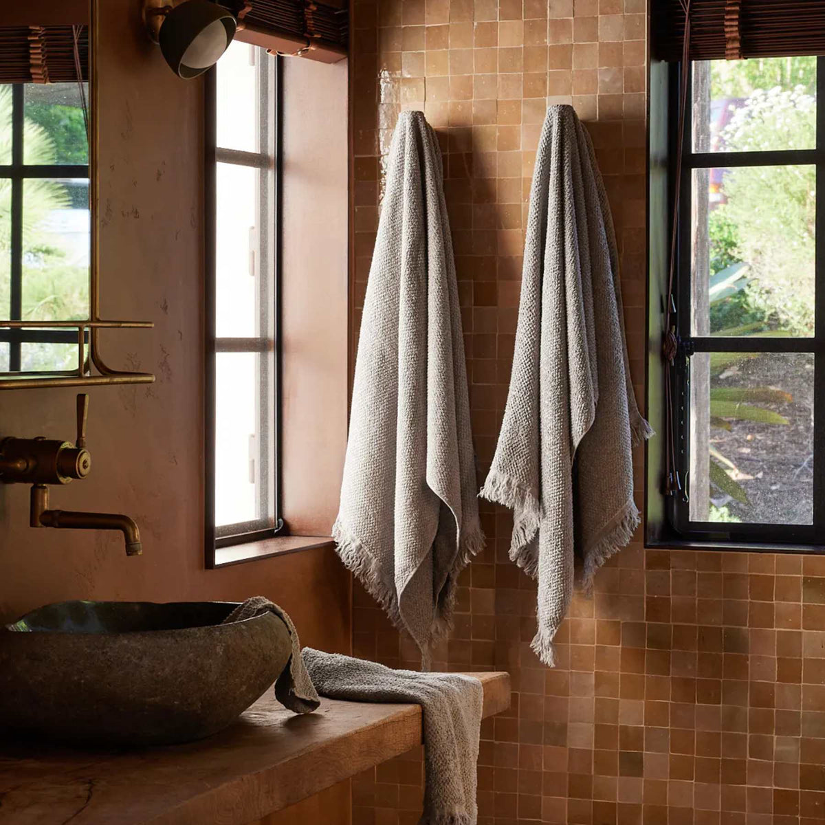 Two beige towels hang on hooks on a tan tiled wall between two windows. A stone sink with a towel draped nearby sits on a wooden countertop, with greenery visible outside.