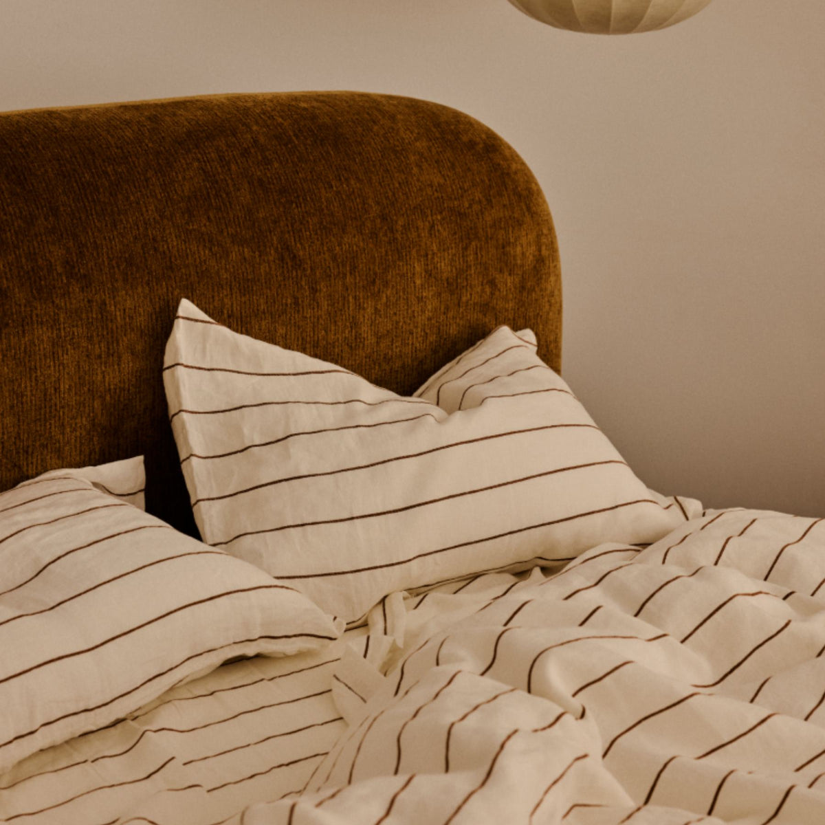A bed with a brown upholstered headboard and white bedding featuring thin black stripes. Two pillows rest against the headboard, and a beige round lampshade hangs above.