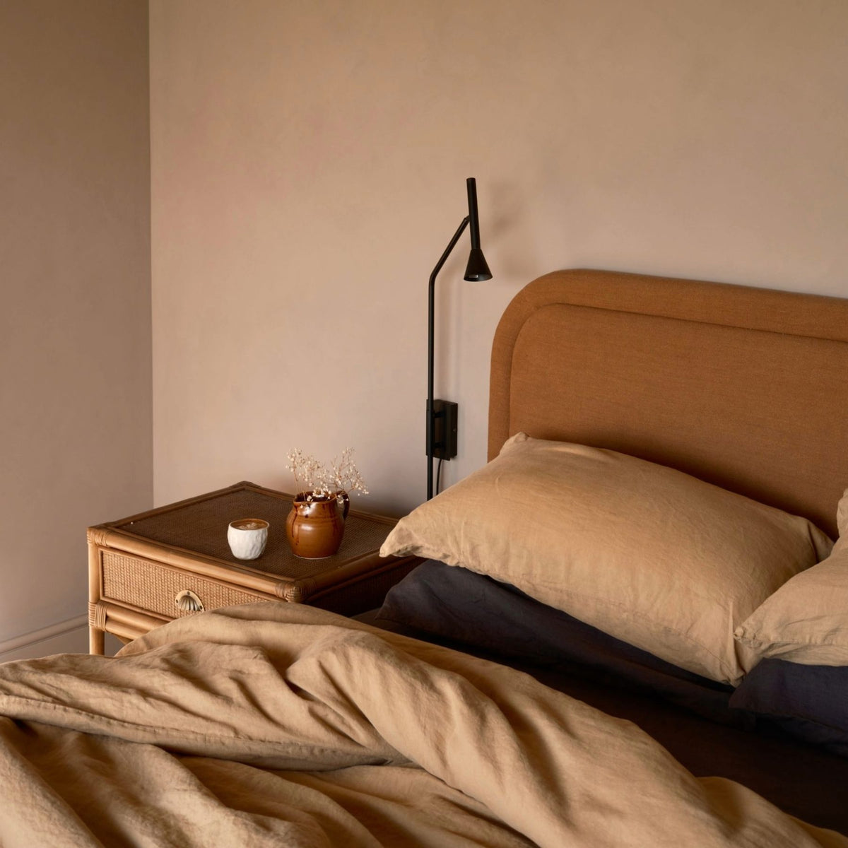 A cozy bed with tan bedding sits beside a wicker nightstand holding a brown vase with dried flowers, a cup, and a book. A black reading lamp is mounted on the wall above the nightstand.