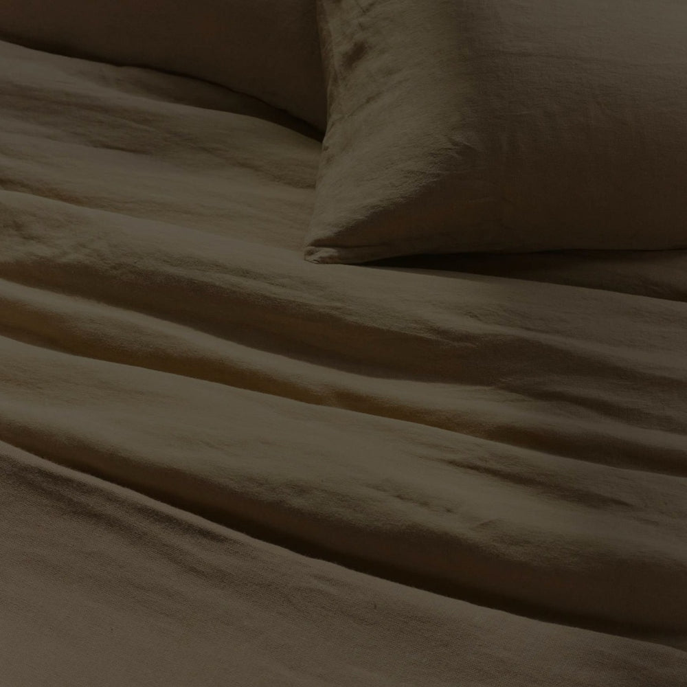 A neatly made bed with beige linen sheets and a pillow, softly wrinkled, viewed in dim lighting.