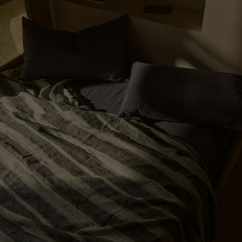 A neatly made bed with two dark pillows and a striped blanket, softly lit by natural light coming from the side.